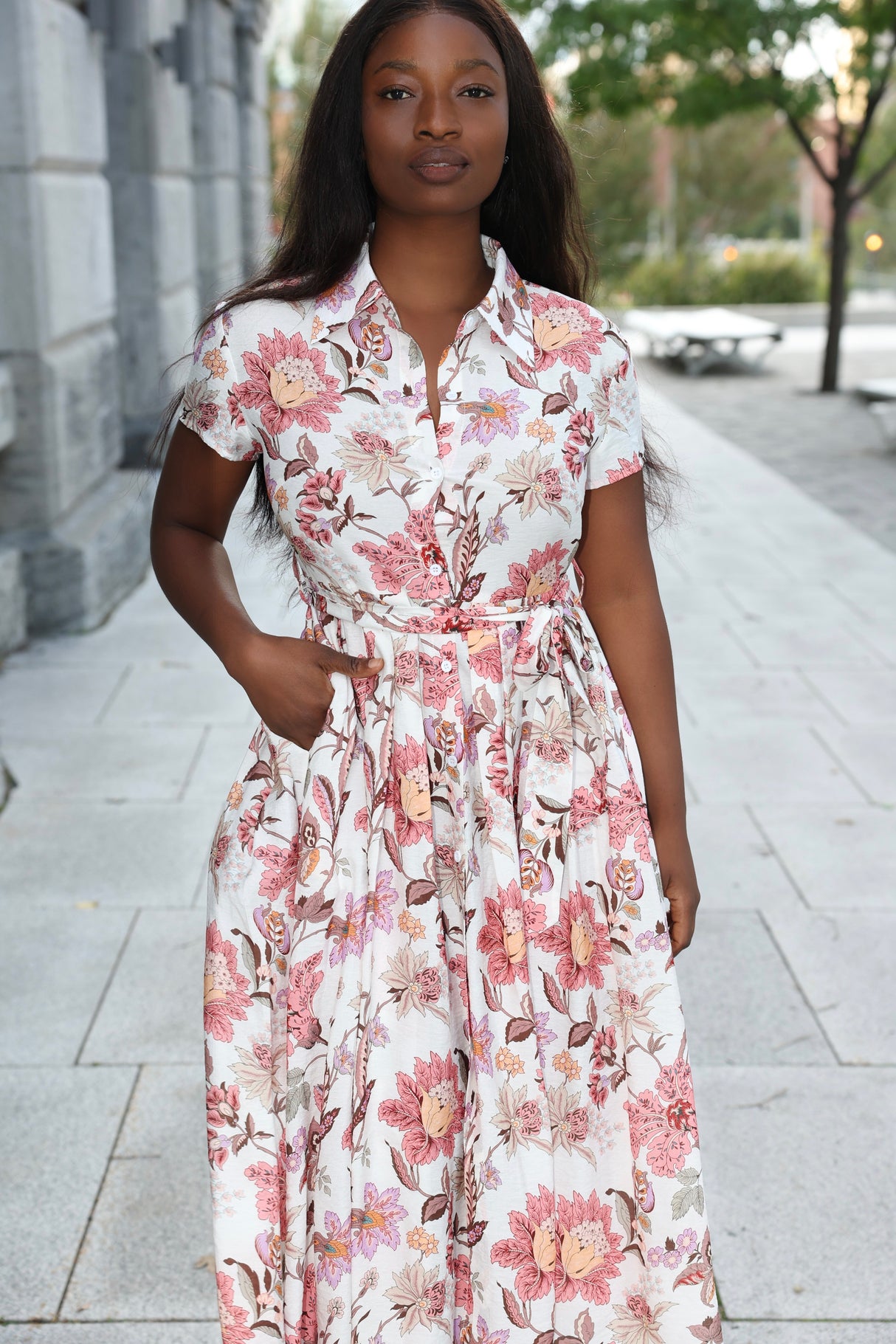 Woman wearing a floral dress standing on a sidewalk with trees and buildings in the background
